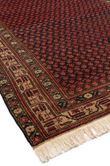 Vista en ángulo de una alfombra con un diseño repetitivo en tonos rojos sobre un fondo oscuro, bordeada por un marco beige decorativo.