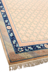 Vista en ángulo de alfombra beige con borde decorativo azul y motivos geométricos suaves.