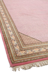 Vista lateral de una alfombra con un diseño rosado, borde marrón y detalles geométricos.