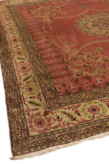 Esquina de una alfombra con bordes decorados en beige y detalles ornamentales en verde y rosa.