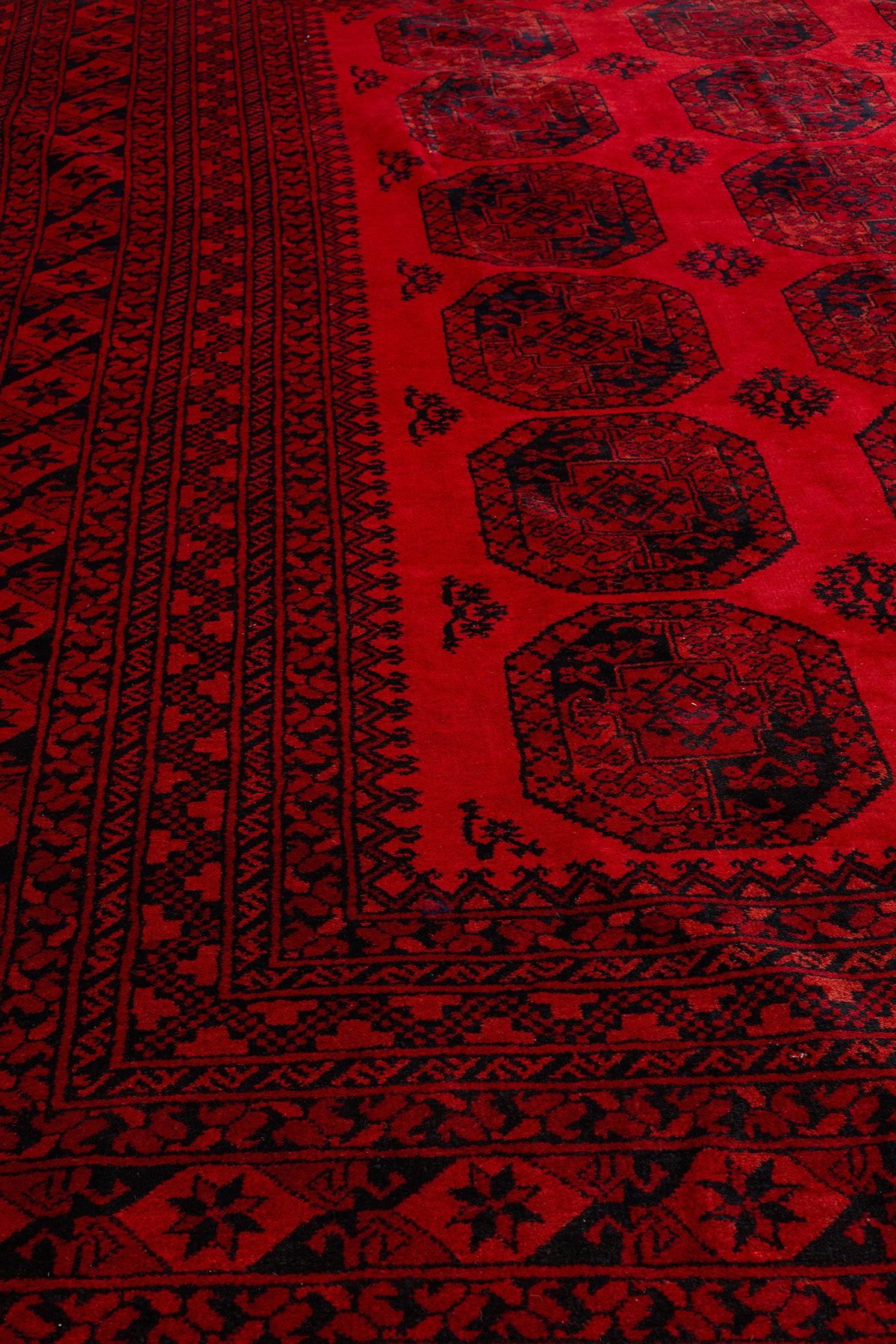 Vista lateral de una alfombra con fondo rojo oscuro, mostrando patrones geométricos y un borde decorado.