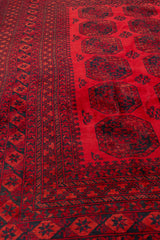 Detalle de la esquina de una alfombra con diseños geométricos en tonos rojo y negro, y flecos claros.