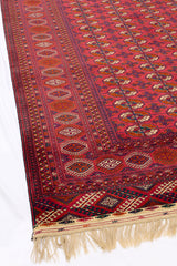 Esquina inferior de una alfombra oriental roja con flecos claros, destacando las figuras geométricas y florales.