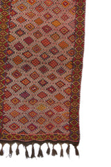 Detalle lateral de alfombra kilim con motivos geométricos sobre fondo burdeos, resaltando sus bordes coloridos.