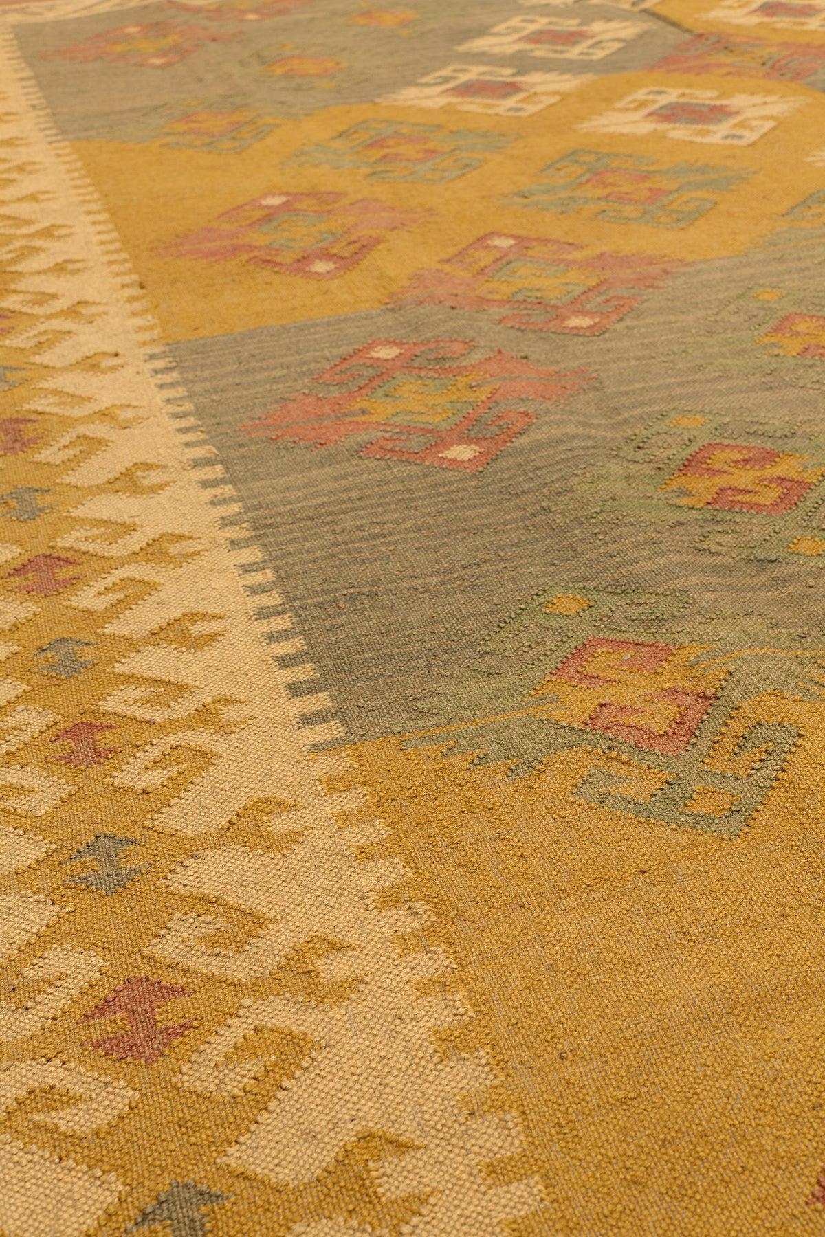 Vista en ángulo de la alfombra kilim, resaltando los detalles de textura y las figuras geométricas sobre un tejido en tonos mostaza y verde.