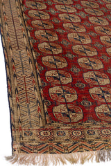 Detalle del borde de una alfombra oriental en tonos beige y azul sobre un fondo rojo.
