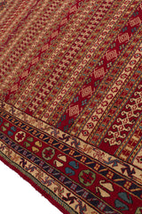 Vista en ángulo de la alfombra resaltando la textura tejida a mano y los patrones simétricos beige sobre fondo rojo.