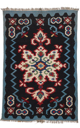 Vista completa de la alfombra con fondo negro y un gran motivo central simétrico de inspiración floral en tonos crema, rojo y verde, enmarcada por una guarda azul con detalles geométricos.