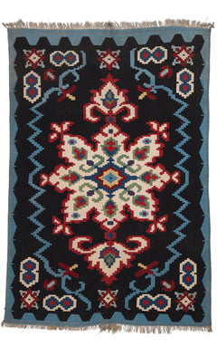 Vista completa de la alfombra con fondo negro y un gran motivo central simétrico de inspiración floral en tonos crema, rojo y verde, enmarcada por una guarda azul con detalles geométricos.