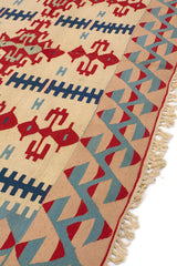 Detalle en perspectiva del borde derecho de la alfombra, con zigzags en rojo y azul sobre fondo beige.