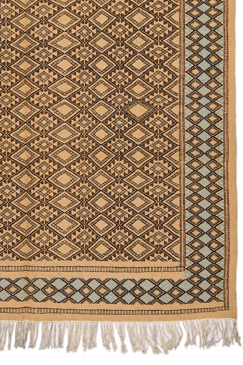 Detalle de la esquina inferior derecha de la alfombra, mostrando el diseño geométrico en diamantes y el borde celeste con flecos beige.
