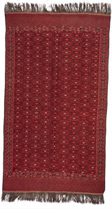 Vista frontal completa de la alfombra roja con patrones geométricos romboidales en tonos oscuros y detalles en beige, bordeada con flecos.