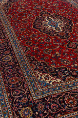 Toma en perspectiva de la alfombra que resalta el brillo de su fondo rojo, el medallón central floral y los bordes con arabescos azul marino y marfil.