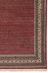Detalle lateral inferior derecho de la alfombra, mostrando el patrón floral rojo y las franjas decoradas con formas geométricas y figuras en tono crema.