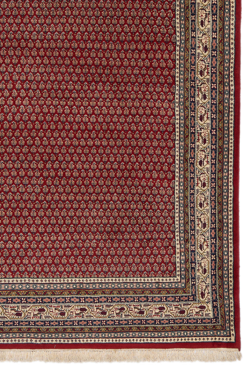 Detalle lateral inferior derecho de la alfombra, mostrando el patrón floral rojo y las franjas decoradas con formas geométricas y figuras en tono crema.