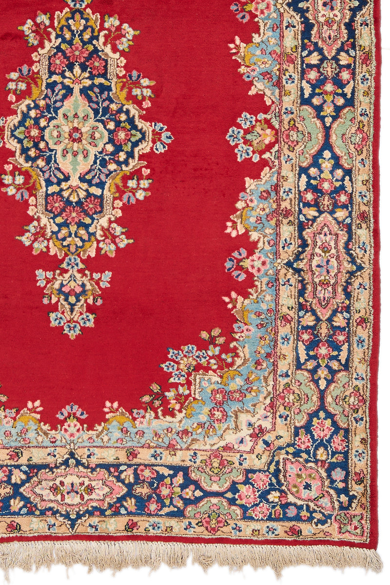 Detalle del medallón central y esquina derecha de la alfombra persa, con intrincados motivos florales sobre fondo rojo y borde azul marino decorado con flores rosadas.