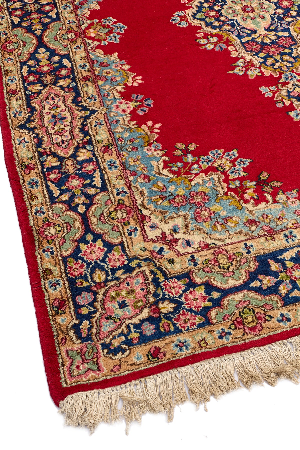 Vista angular del borde inferior izquierdo de la alfombra persa, mostrando la transición entre el campo rojo y la guardas florales en beige, azul, verde y rosado.