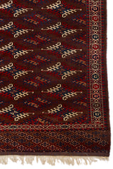 Detalle del borde derecho de la alfombra mostrando patrones geométricos simétricos en rojo, azul y beige sobre un fondo marrón intenso.