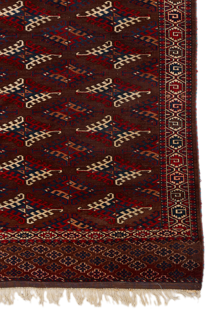 Detalle del borde derecho de la alfombra mostrando patrones geométricos simétricos en rojo, azul y beige sobre un fondo marrón intenso.