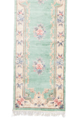 Detalle inferior de la alfombra mostrando las flores rosadas y azules enmarcadas en un borde beige con diseño ornamental.