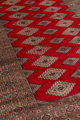 Perspectiva angular de la alfombra destacando el diseño repetitivo de rombos sobre fondo rojo intenso y la guarda beige.