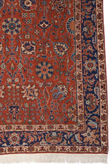 Detalle del borde de una alfombra roja con motivos florales y guarda azul decorativa, mostrando la artesanía y los colores vibrantes.