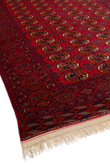 Vista lateral de una alfombra oriental roja con un diseño geométrico intrincado y flecos decorativos en los bordes.