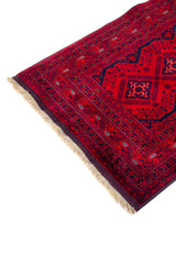 Detalle en perspectiva de alfombra roja con borde geométrico, motivos decorativos en azul oscuro y flecos beige en el extremo.