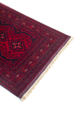 Vista cercana de una esquina de alfombra oriental roja, con patrones geométricos, borde azul oscuro y flecos claros.