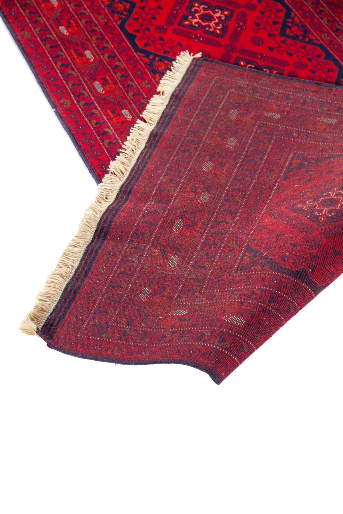 Detalle de alfombra roja doblada que muestra el reverso tejido, flecos claros y diseño geométrico tradicional en tonos oscuros.