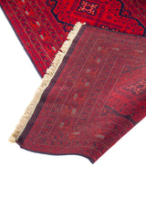 Detalle de alfombra roja doblada que muestra el reverso tejido, flecos claros y diseño geométrico tradicional en tonos oscuros.