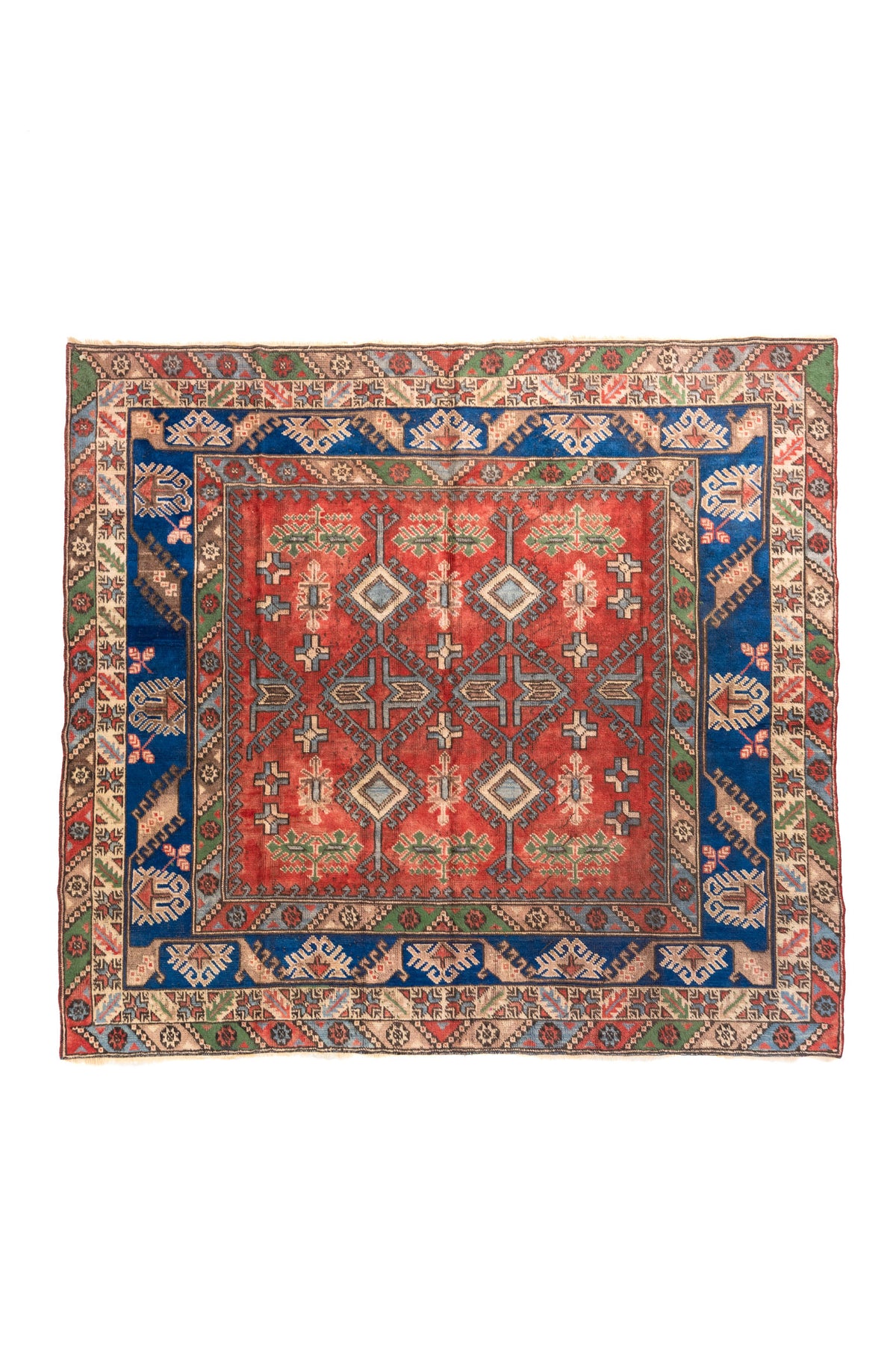 Alfombra estilo Kazak en formato casi cuadrado, con fondo rojo, guarda azul intensa y motivos geométricos tribales en tonos blanco suave, verde y celeste.