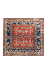 Alfombra estilo Kazak en formato casi cuadrado, con fondo rojo, guarda azul intensa y motivos geométricos tribales en tonos blanco suave, verde y celeste.