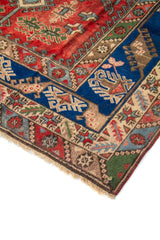 Primer plano de borde y esquina de alfombra oriental en tonos rojo, azul y beige, con patrones geométricos tradicionales y detalles multicolor.