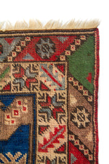 Acercamiento de esquina de alfombra tejida con motivos geométricos pequeños, borde decorativo y contraste entre rojo, azul y verde.
