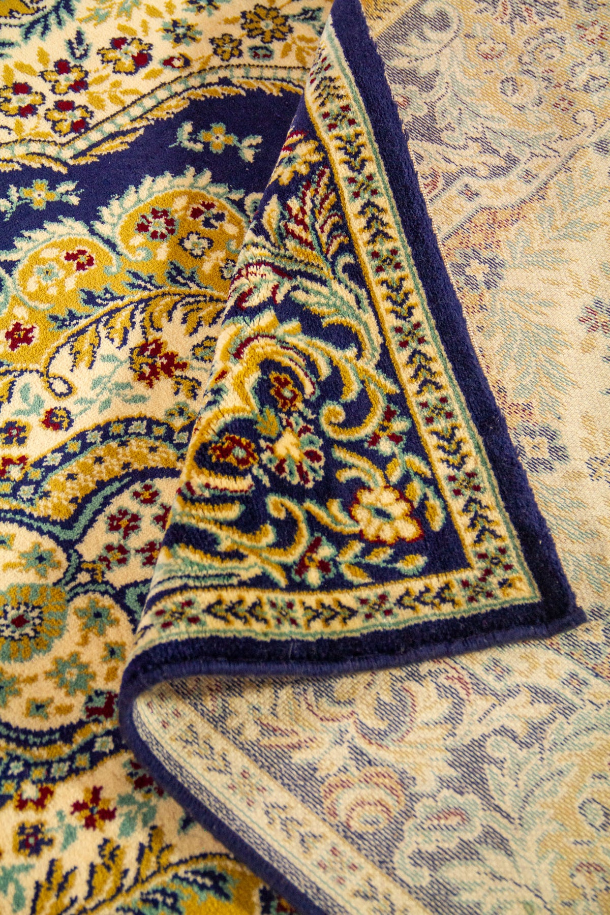 Alfombra parcialmente enrollada que muestra la textura, el reverso tejido y el diseño ornamental floral en tonos azul marino, beige y dorado.
