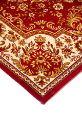 Primer plano de borde y esquina de alfombra clásica en tonos rojo, blanco suave y dorado, con motivos florales curvos y acabado prolijo.