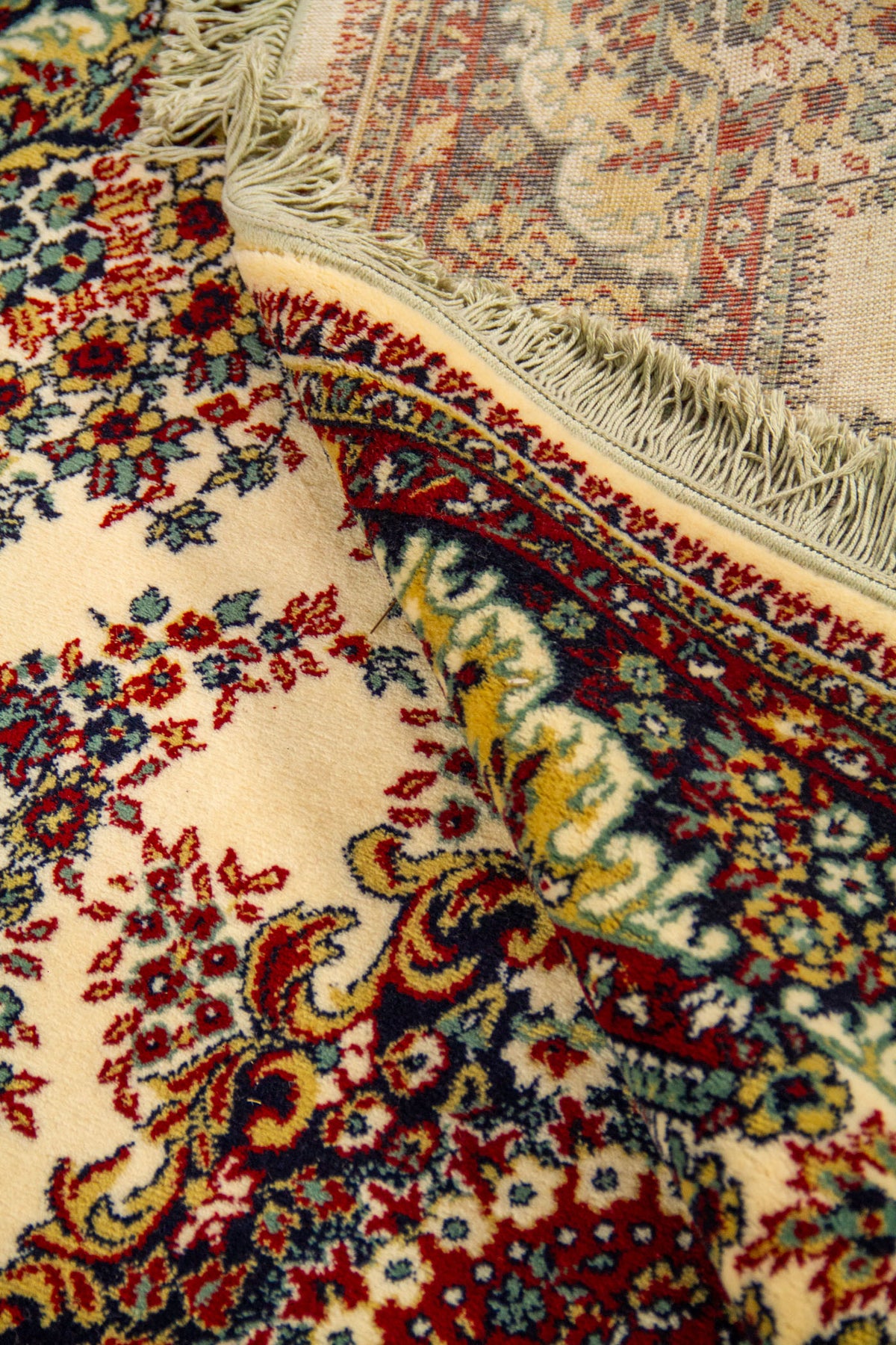 Detalle de alfombra doblada que deja ver el reverso, los flecos y la densidad del diseño floral central sobre fondo blanco suave.