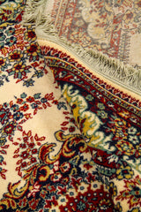 Detalle de alfombra doblada que deja ver el reverso, los flecos y la densidad del diseño floral central sobre fondo blanco suave.