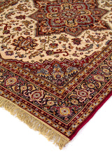 Detalle de esquina de alfombra oriental con flecos beige, campo beige, medallón geométrico central y guarda floral en rojo, negro y dorado.