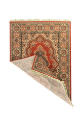 Alfombra clásica color rojo con una esquina doblada que deja ver el reverso, medallón central elaborado, borde decorativo con motivos florales repetidos y flecos beige.