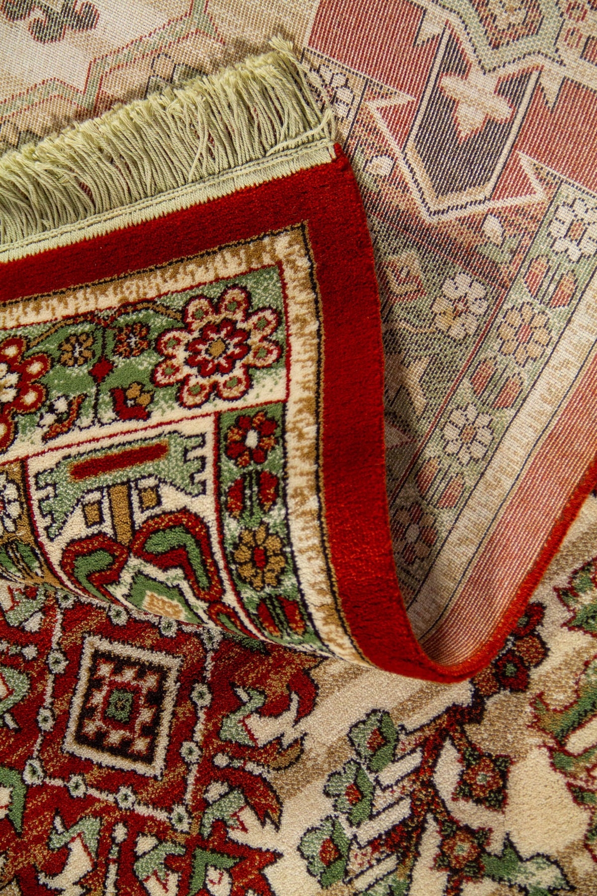 Detalle de alfombra doblada mostrando el reverso tejido, el canto rojo aterciopelado y los flecos color blanco suave.