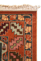 Primer plano de la esquina superior de alfombra geométrica con borde rojo, motivos simétricos y flecos color beige claro.