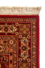 Primer plano de la esquina de una alfombra roja clásica con flecos beige, guarda floral y terminación de borde aterciopelado.