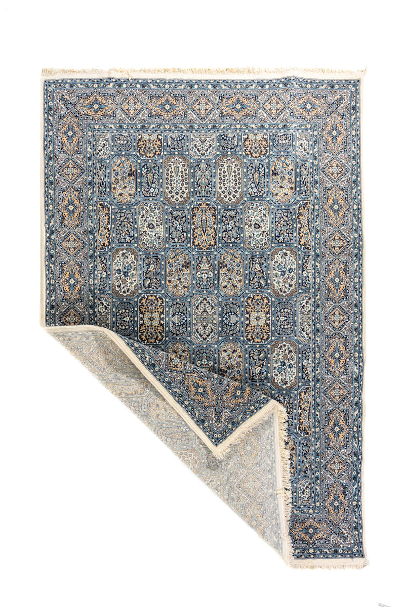 Alfombra azul y beige con esquina doblada que muestra el reverso, los flecos y el diseño de paneles ornamentales