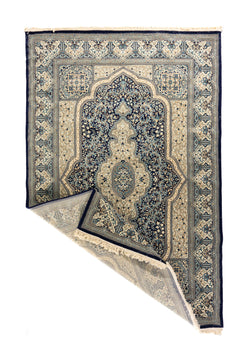 Alfombra azul y beige con esquina doblada que muestra el reverso, los flecos y el diseño central decorativo