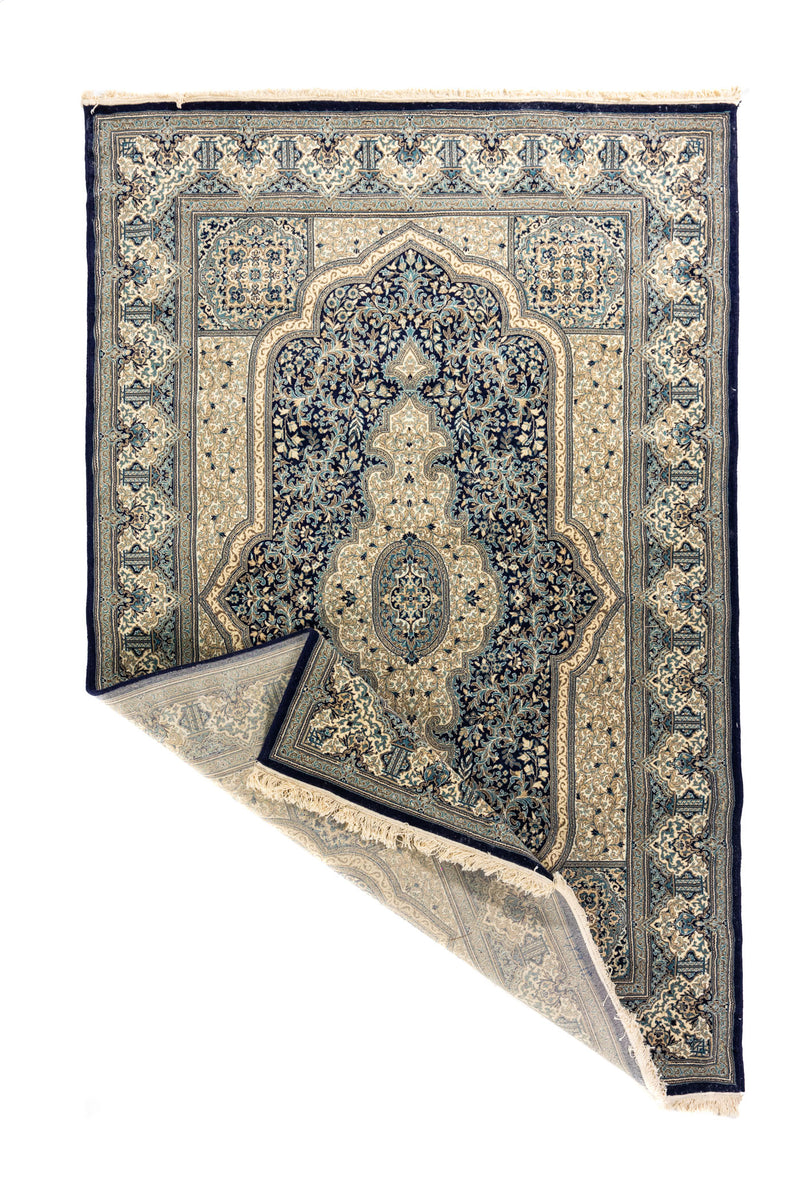 Alfombra azul y beige con esquina doblada que muestra el reverso, los flecos y el diseño central decorativo