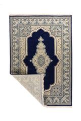 Alfombra clásica azul y blanco suave con una esquina doblada, mostrando el reverso, los flecos claros y la densidad del tejido decorativo.