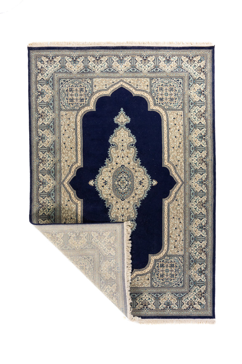 Alfombra clásica azul y blanco suave con una esquina doblada, mostrando el reverso, los flecos claros y la densidad del tejido decorativo.