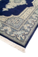 Detalle en perspectiva de esquina de alfombra oriental con fondo azul profundo, borde ornamental en blanco suave y celeste, y flecos en el extremo.